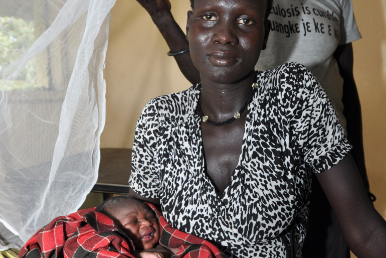 Eine Mutter hält erschöpft aber glücklich ihr Neugeborenes in den Armen. Die traditionelle Geburtshelferin im Hintergrund half ihr bei der Entbindung in der von Hoffnungszeichen betriebenen Buschklinik in Nyal/Südsudan.