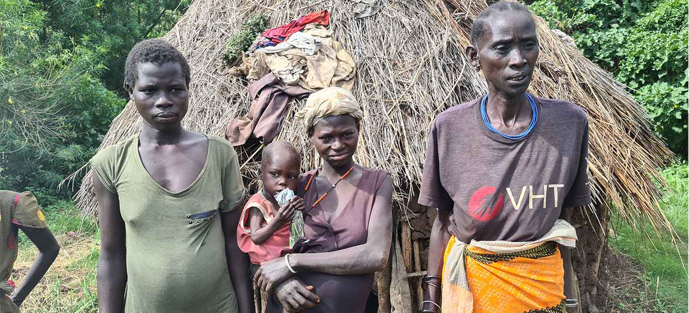 Drei Frauen stehen vor einer Hütte, die Frau in der Mitte hält ein Baby auf dem Arm