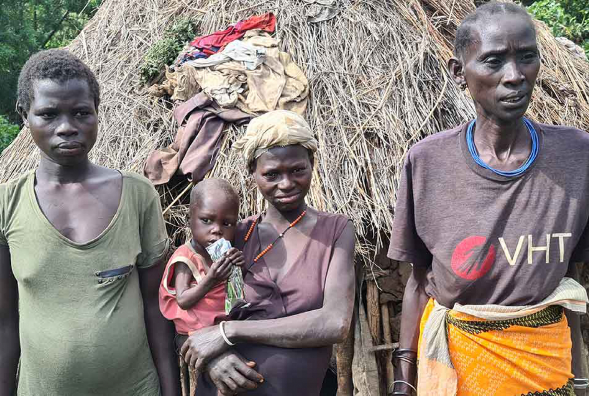 Drei Frauen stehen vor einer Hütte, die Frau in der Mitte hält ein Baby auf dem Arm